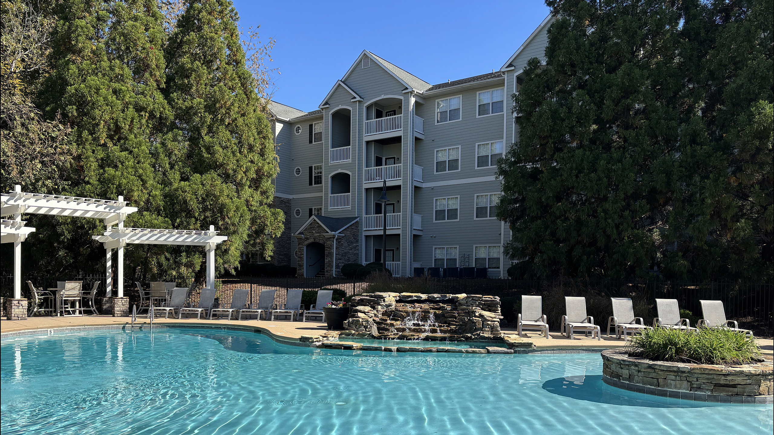 Inviting outdoor pool area featuring elegant lounge chairs and pergola, set against a modern apartment building with lush greenery.