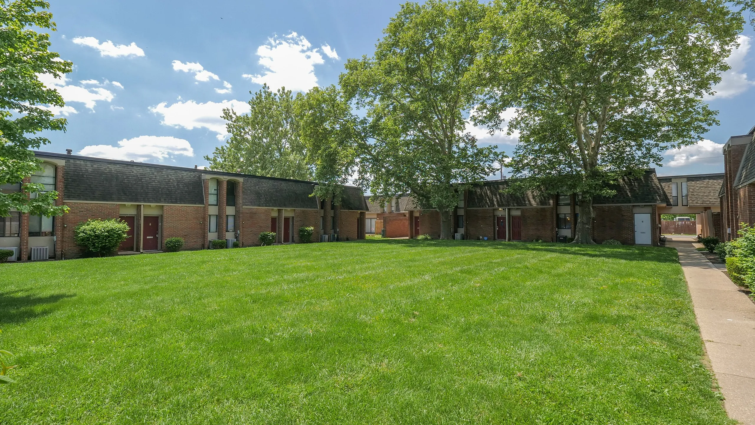 Charming apartment community with brick buildings, lush green lawns, and mature trees under a clear blue sky.