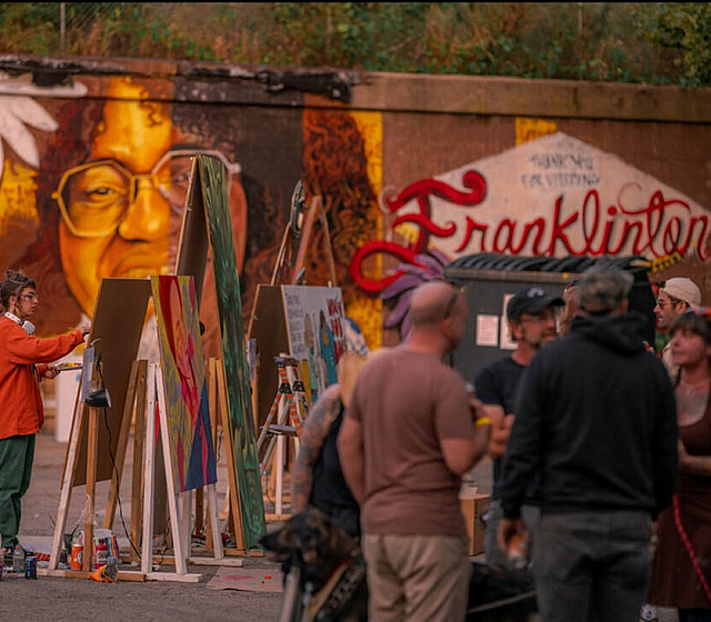 Vibrant outdoor art scene with artists painting on easels in front of colorful murals, creating a lively community atmosphere.