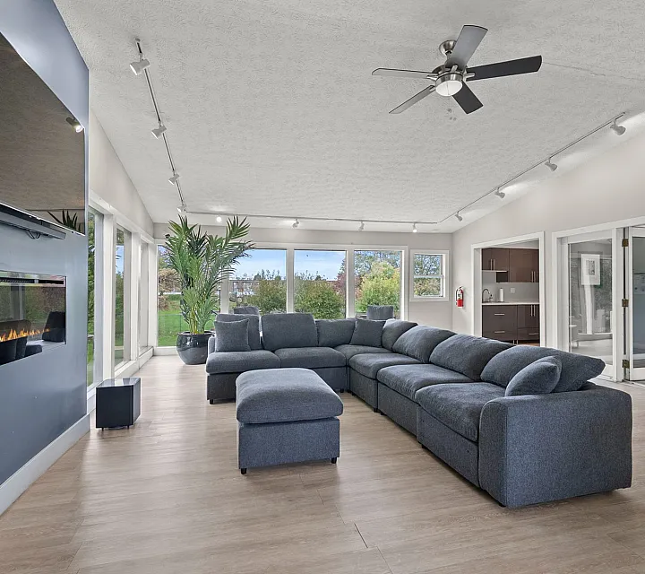 Elegant living room with modern gray sectional, large windows revealing green views, sleek wall-mounted fireplace, and stylish ceiling fan.