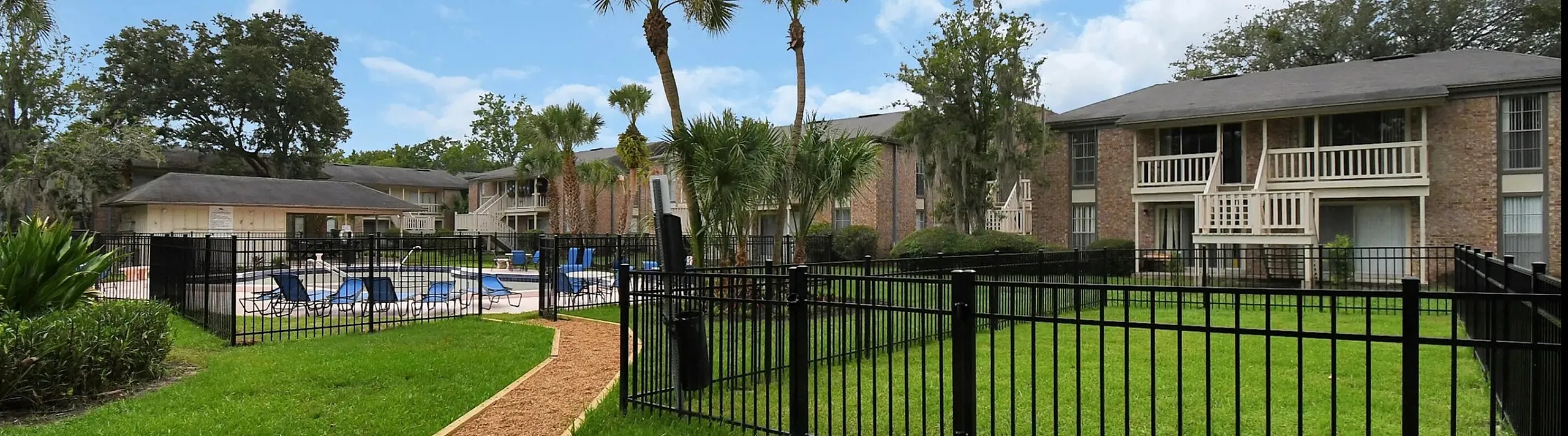 Charming apartment complex with lush green lawns, a welcoming walkway, and a refreshing pool under a bright blue sky.