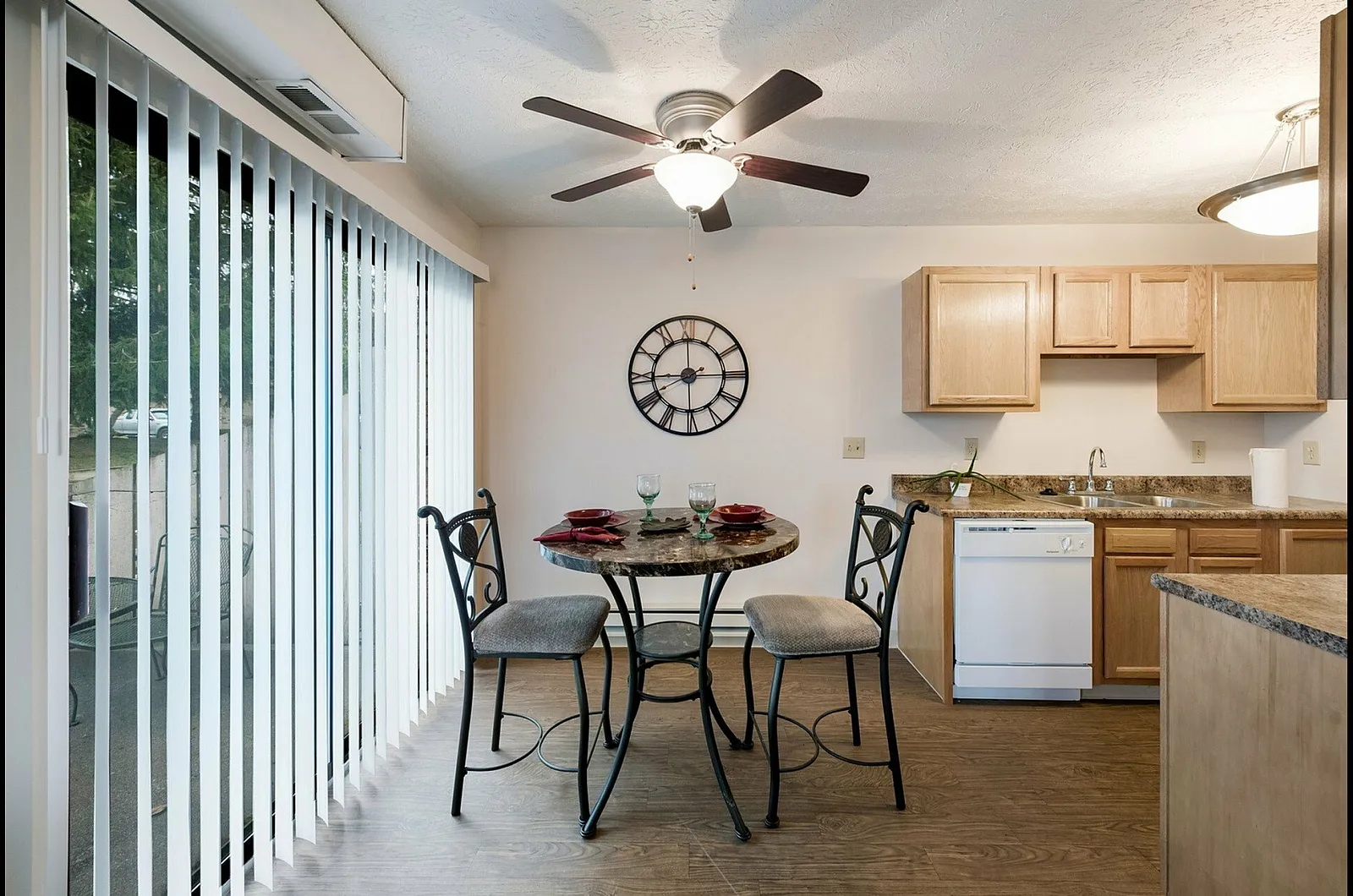 Charming dining area with a round table set for two, large windows with vertical blinds, and a modern kitchen with wooden cabinetry and sleek countertops.