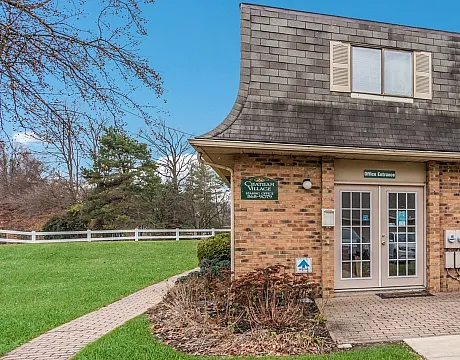 Charming two-story brick building with a unique Mansard roof, set in a lush green area, offering a welcoming office entrance.