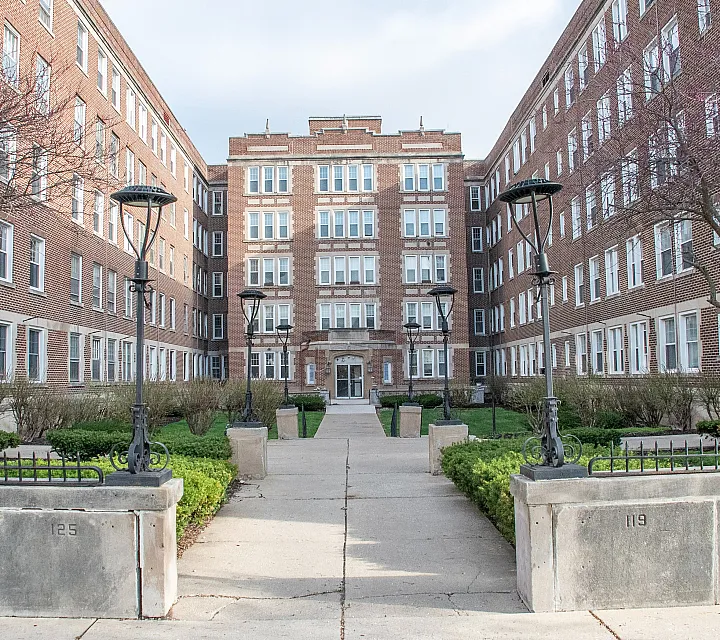 Charming brick apartment building with an inviting courtyard, highlighted by lush greenery and elegant lampposts, perfect for a peaceful urban living experience.
