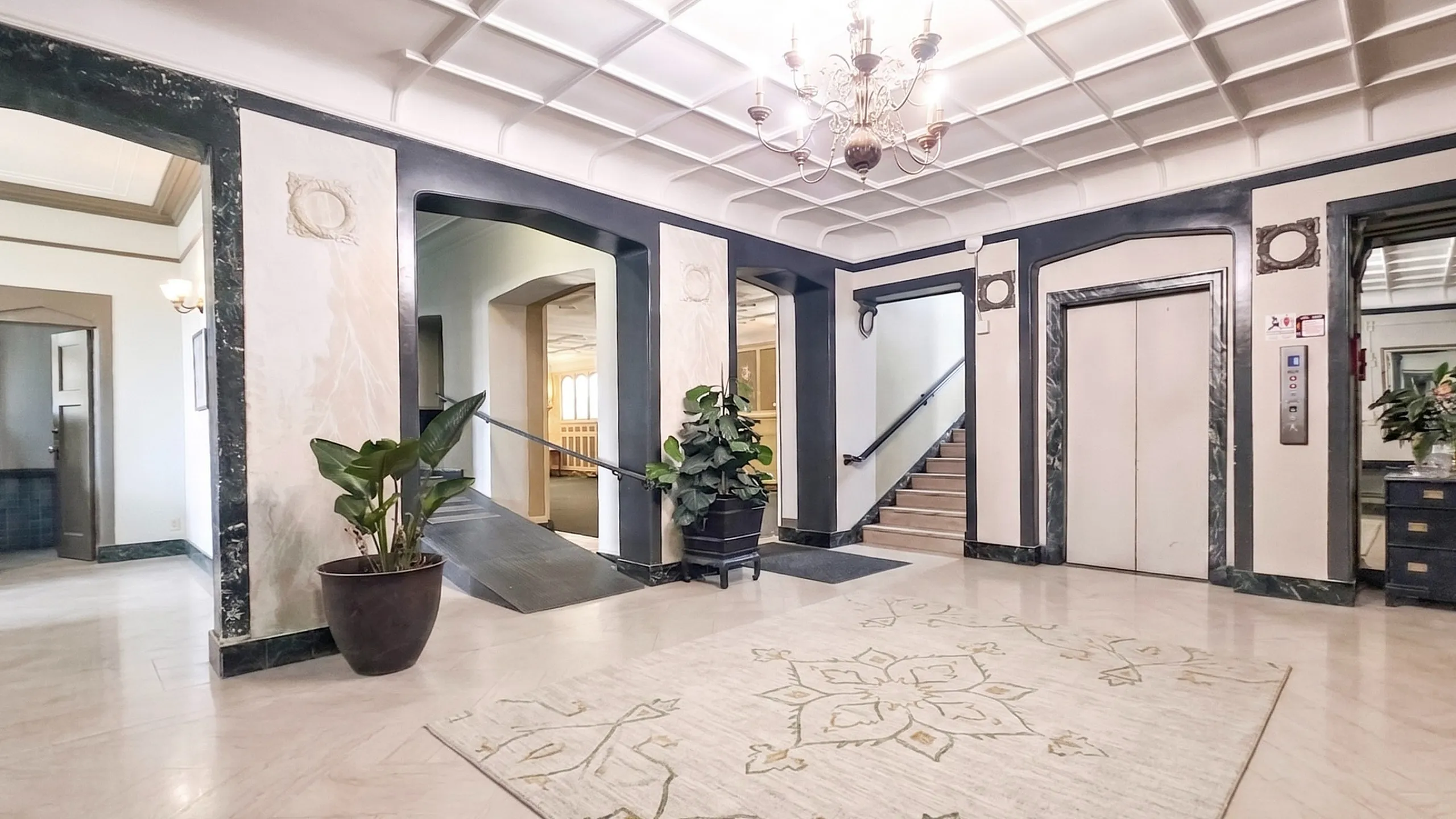 Elegant apartment lobby with classic chandelier, stylish marble accents, and lush greenery, offering an inviting atmosphere.