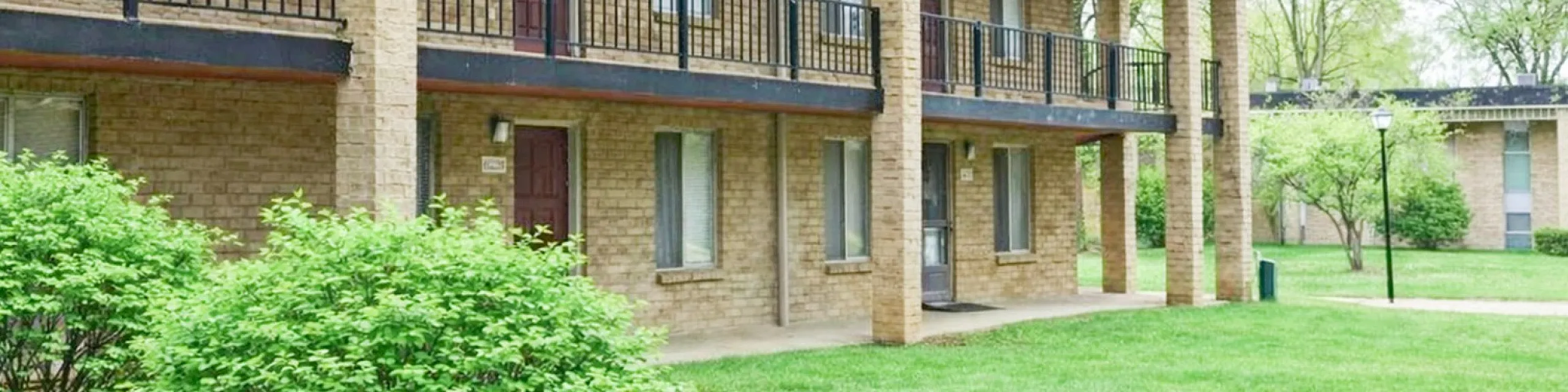 Charming apartment exterior with brick facade, spacious balconies, and lush green landscaping.