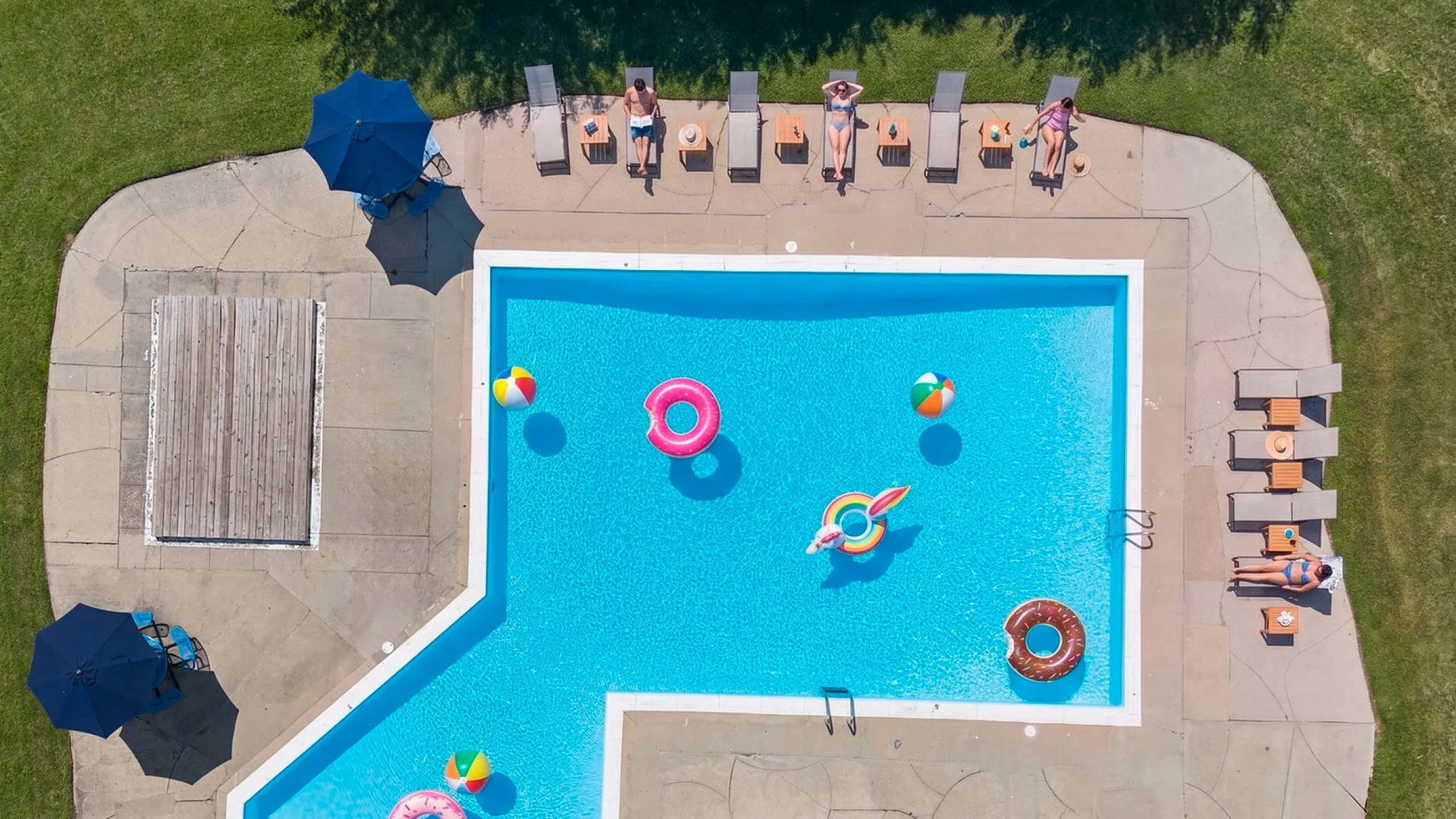 Inviting aerial view of an L-shaped outdoor pool with vibrant inflatables, surrounded by sun loungers and umbrellas on a sunny day.