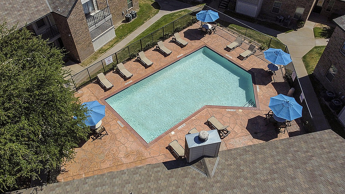 Aerial view of an outdoor swimming pool surrounded by lounge chairs and blue umbrellas, set within an apartment community