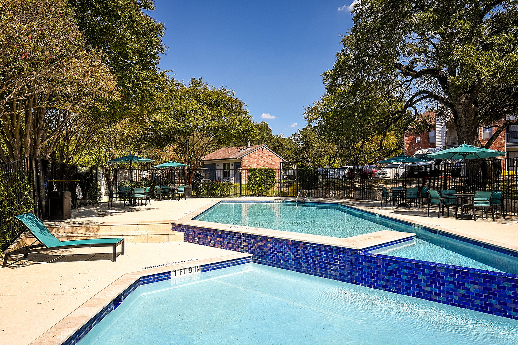 Inviting outdoor pool area with vibrant blue tiles, surrounded by lush trees and patio seating under teal umbrellas, perfect for relaxation and summer gatherings.