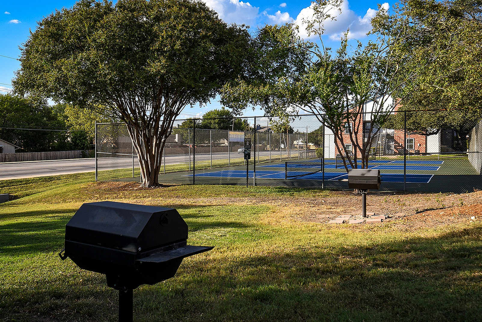 Charming outdoor area with lush green grass, trees, a barbecue grill, and a well-maintained tennis court under a bright blue sky.