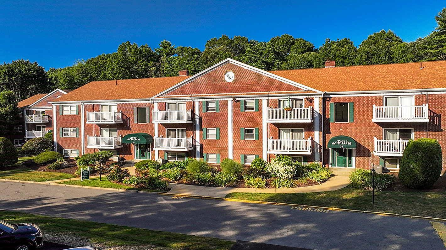 Charming brick apartment complex with white balconies, surrounded by lush greenery and a bright blue sky.