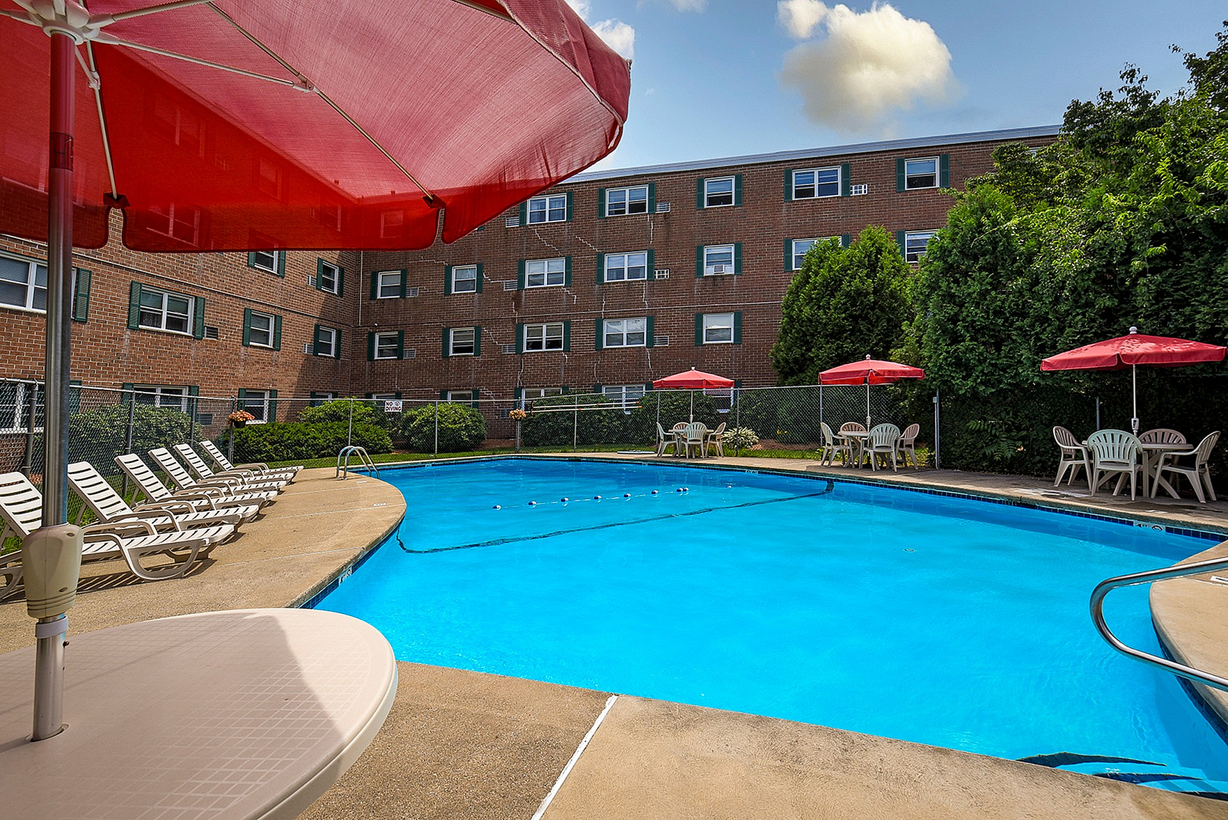 Charming apartment complex with a sparkling outdoor pool, comfortable lounge chairs, and vibrant red umbrellas creating a perfect relaxation spot.