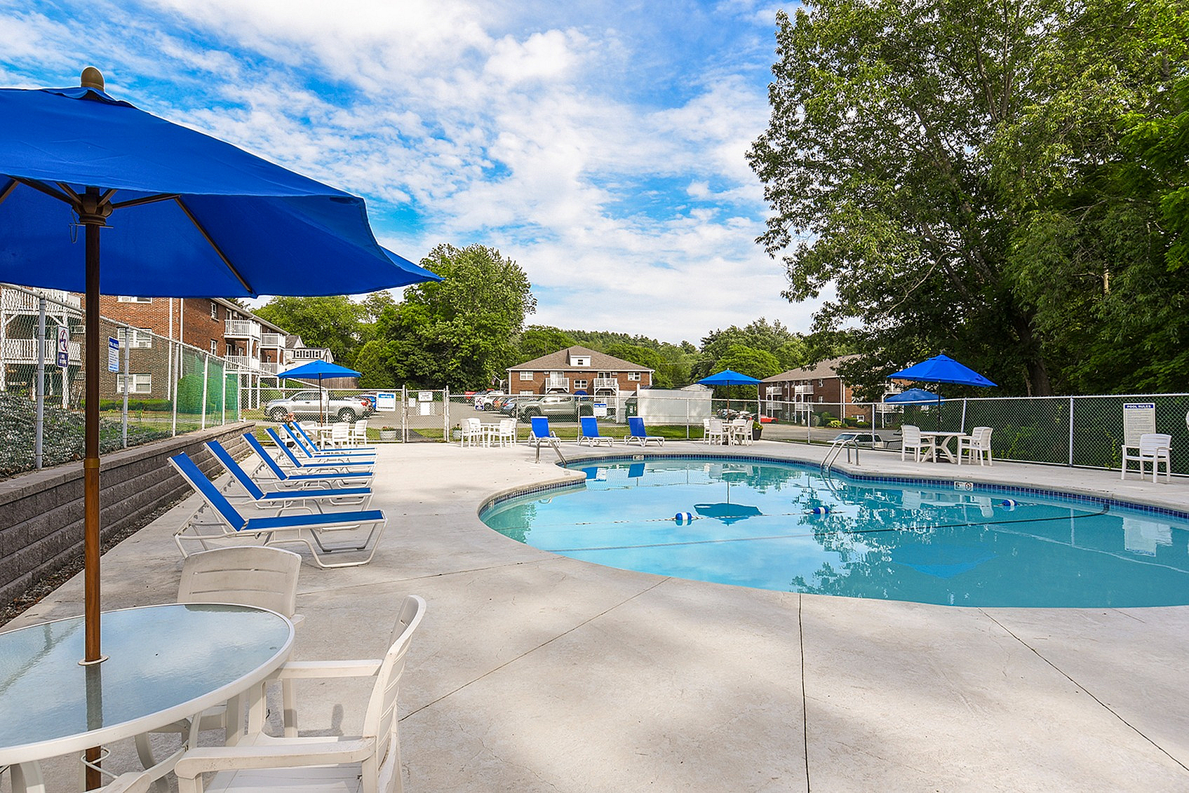 Inviting outdoor pool area with blue umbrellas, sun loungers, and tables, surrounded by lush greenery and apartment buildings.
