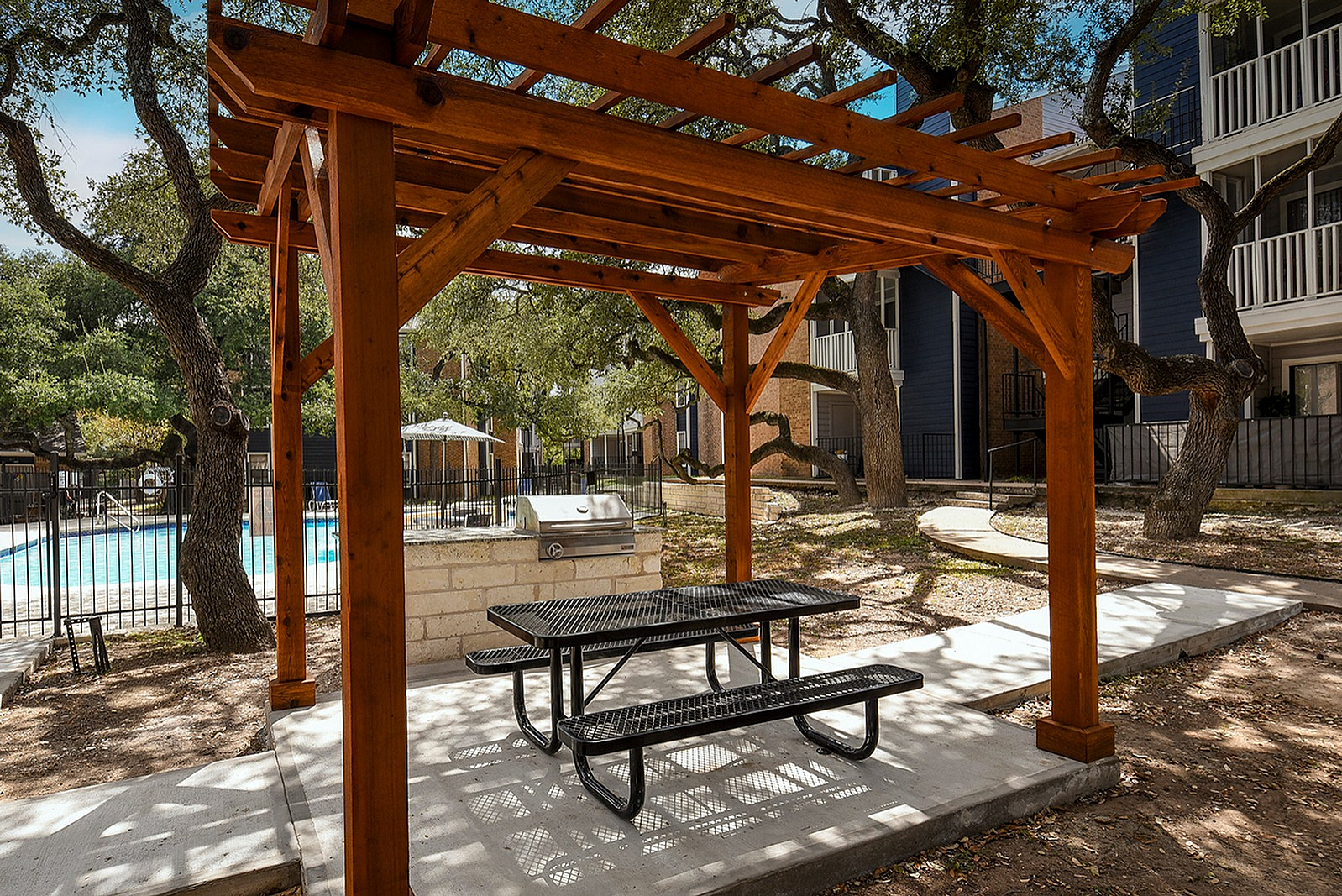 Charming outdoor space with a wooden pergola, picnic table, and grill set against a serene poolside backdrop.