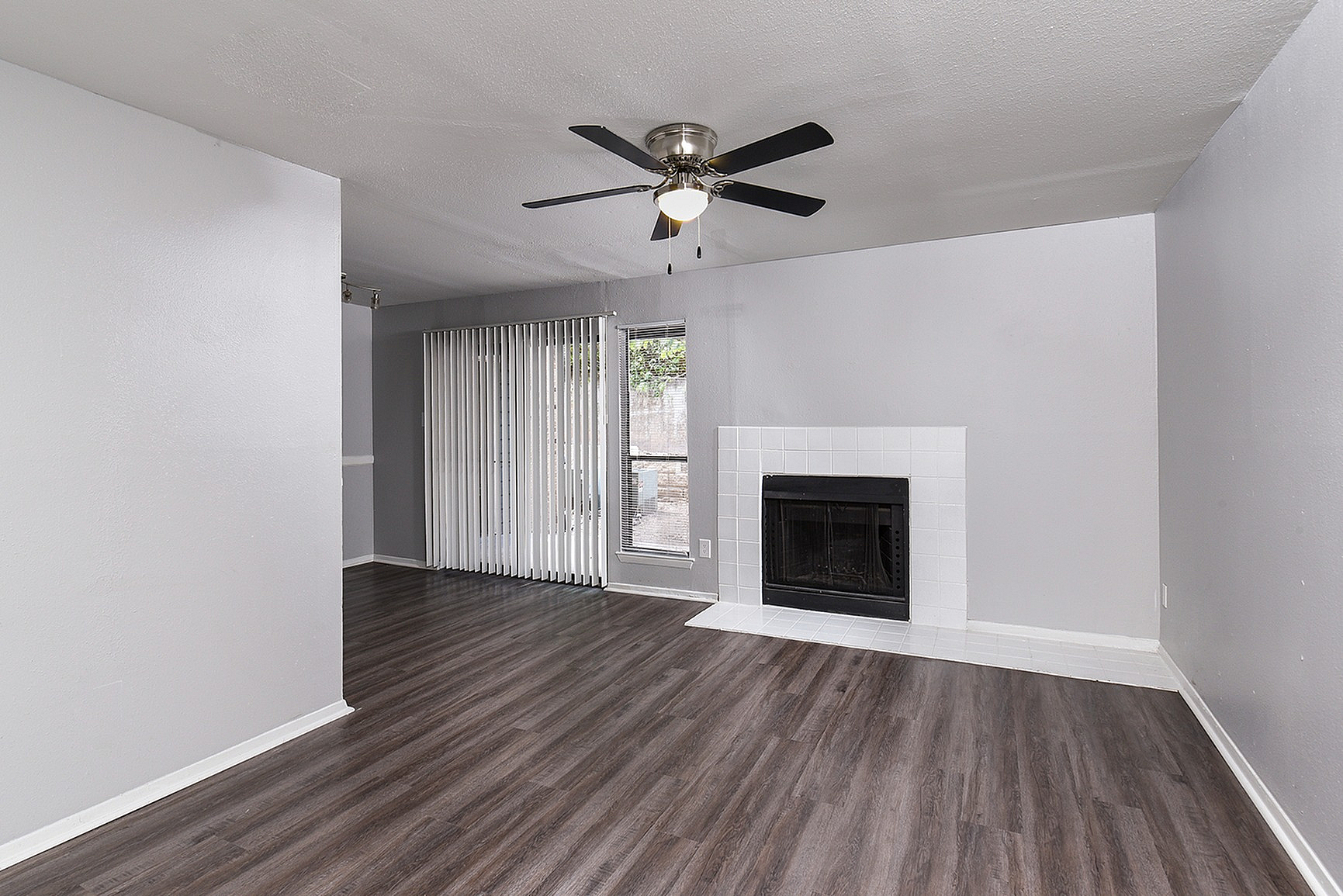 Modern living room with elegant wood flooring, a sleek ceiling fan, and a cozy fireplace, complemented by sunlit vertical blinds.