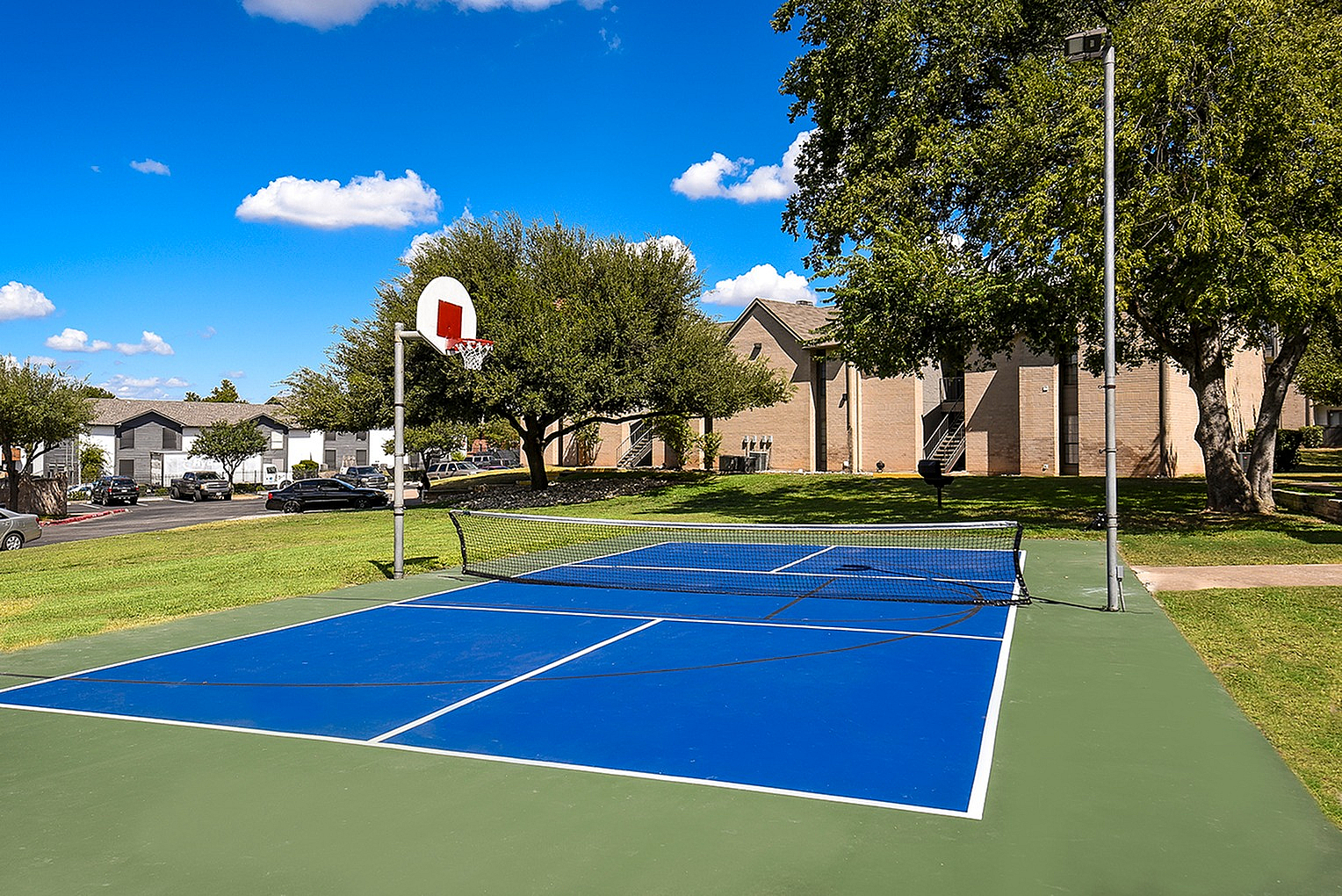 Inviting apartment complex with a vibrant blue sport court, perfect for both tennis and basketball, surrounded by lush green lawns and trees under a clear blue sky.