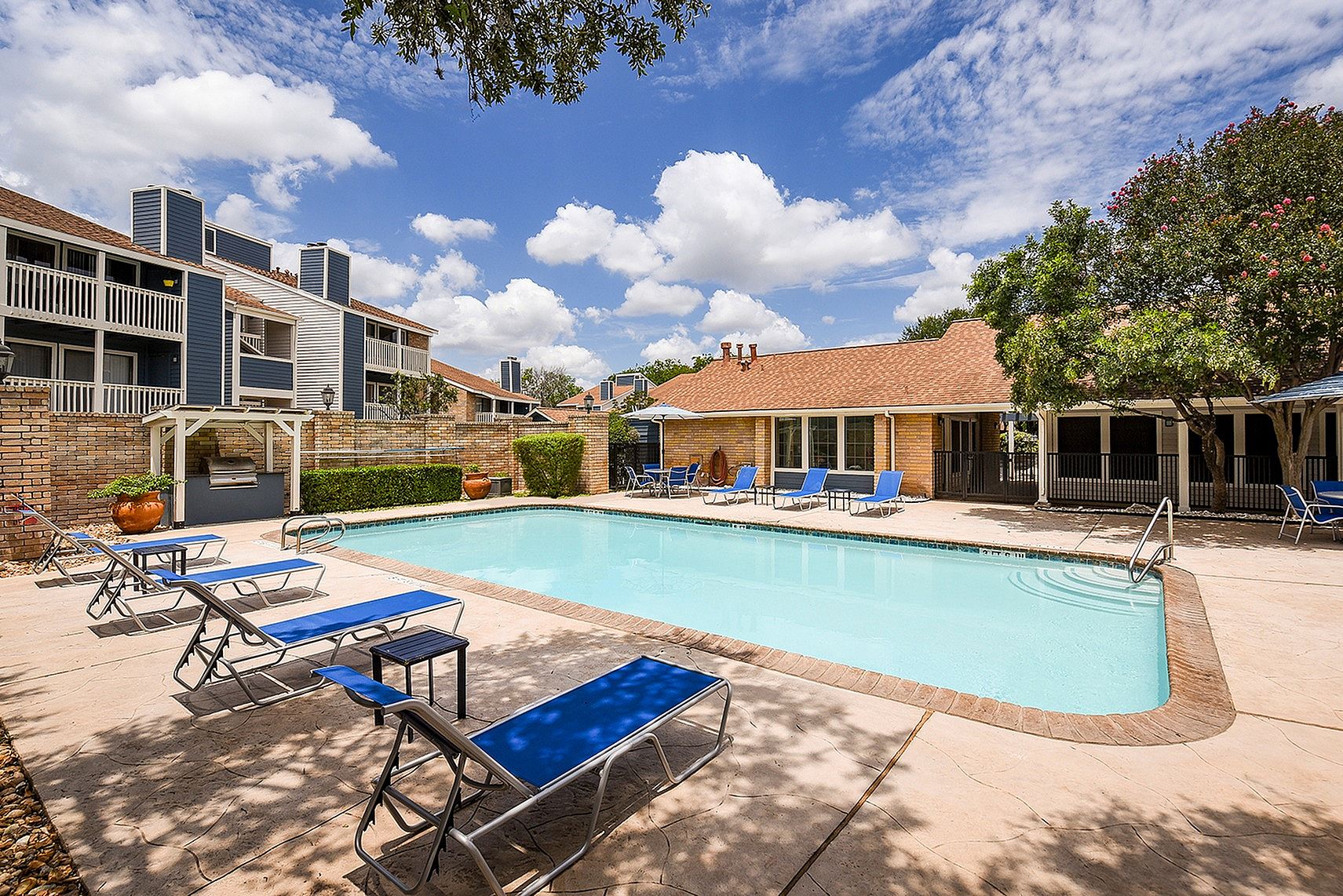 Inviting apartment complex pool area with sun loungers, surrounded by lush greenery and modern buildings under a clear blue sky.