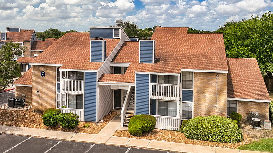 Charming two-story apartment building with red sloped roofs, private balconies, and surrounded by lush greenery under a bright blue sky with fluffy clouds.