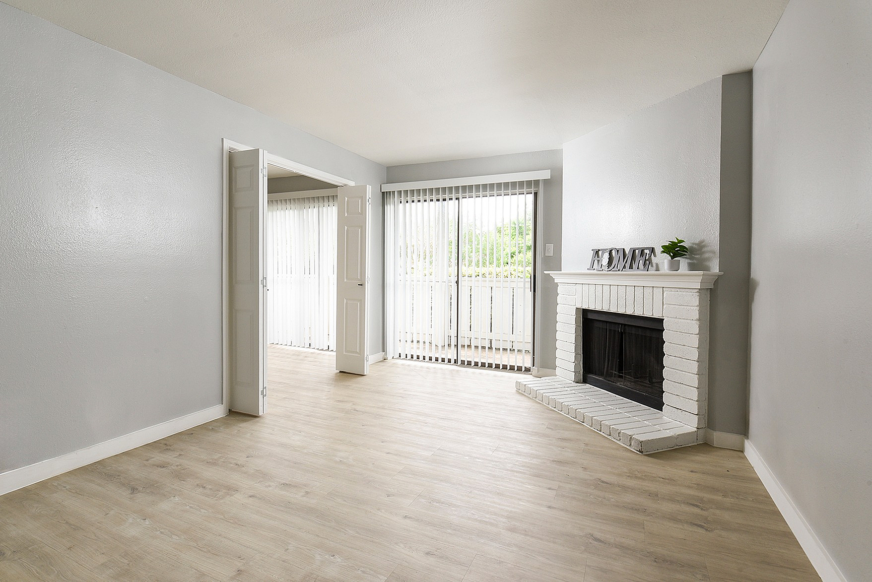 Bright and airy living room with elegant gray walls, a cozy white brick fireplace, and expansive sliding glass doors opening to a private balcony.