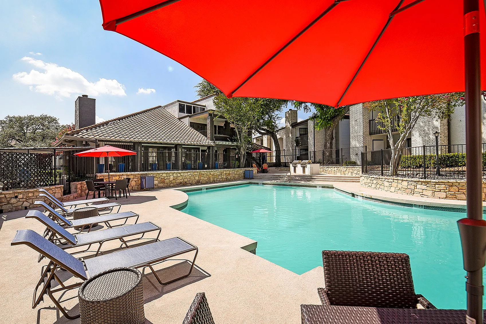 Inviting apartment pool area with stylish loungers and vibrant red umbrellas, perfect for relaxation and sunbathing.