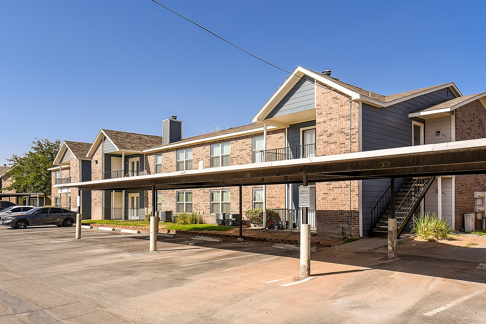 Charming two-story apartment complex with modern brick facade, covered parking, and lush greenery under a clear blue sky.