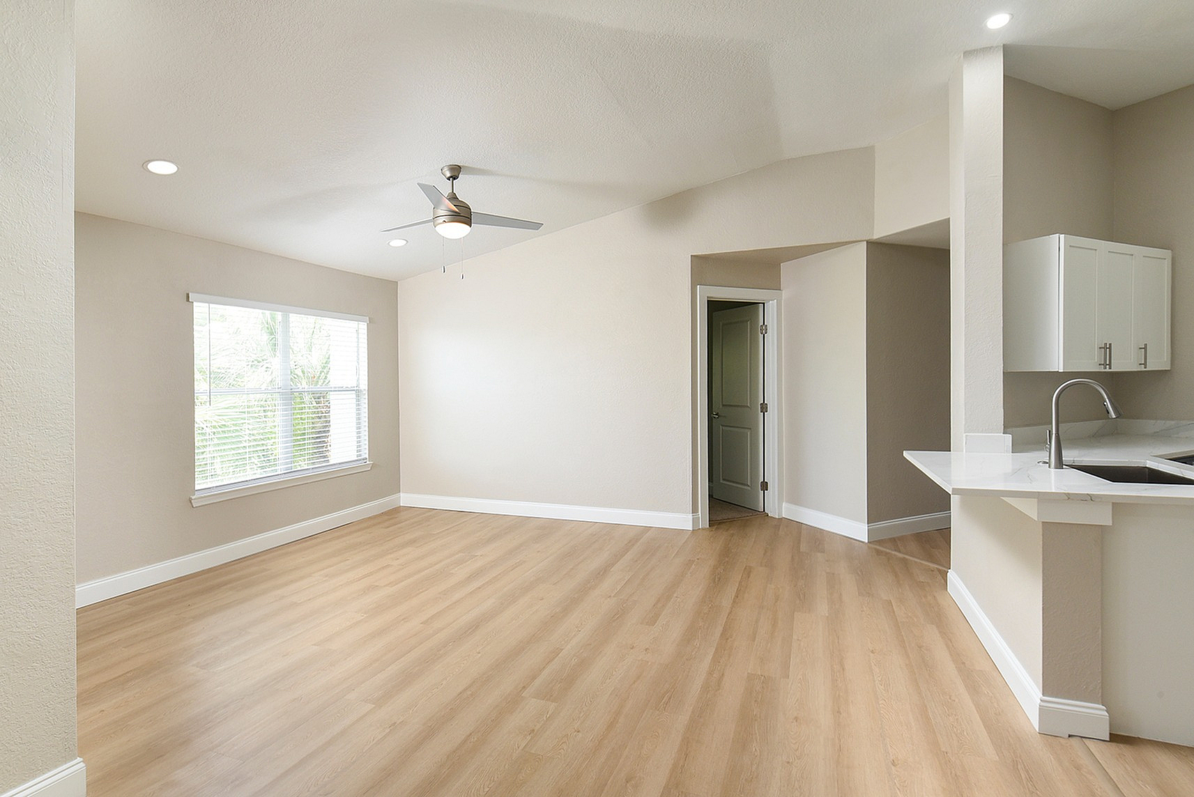 Bright and airy living space with modern wood flooring, neutral tones, and a sleek ceiling fan, adjacent to a chic kitchen with a marble countertop.