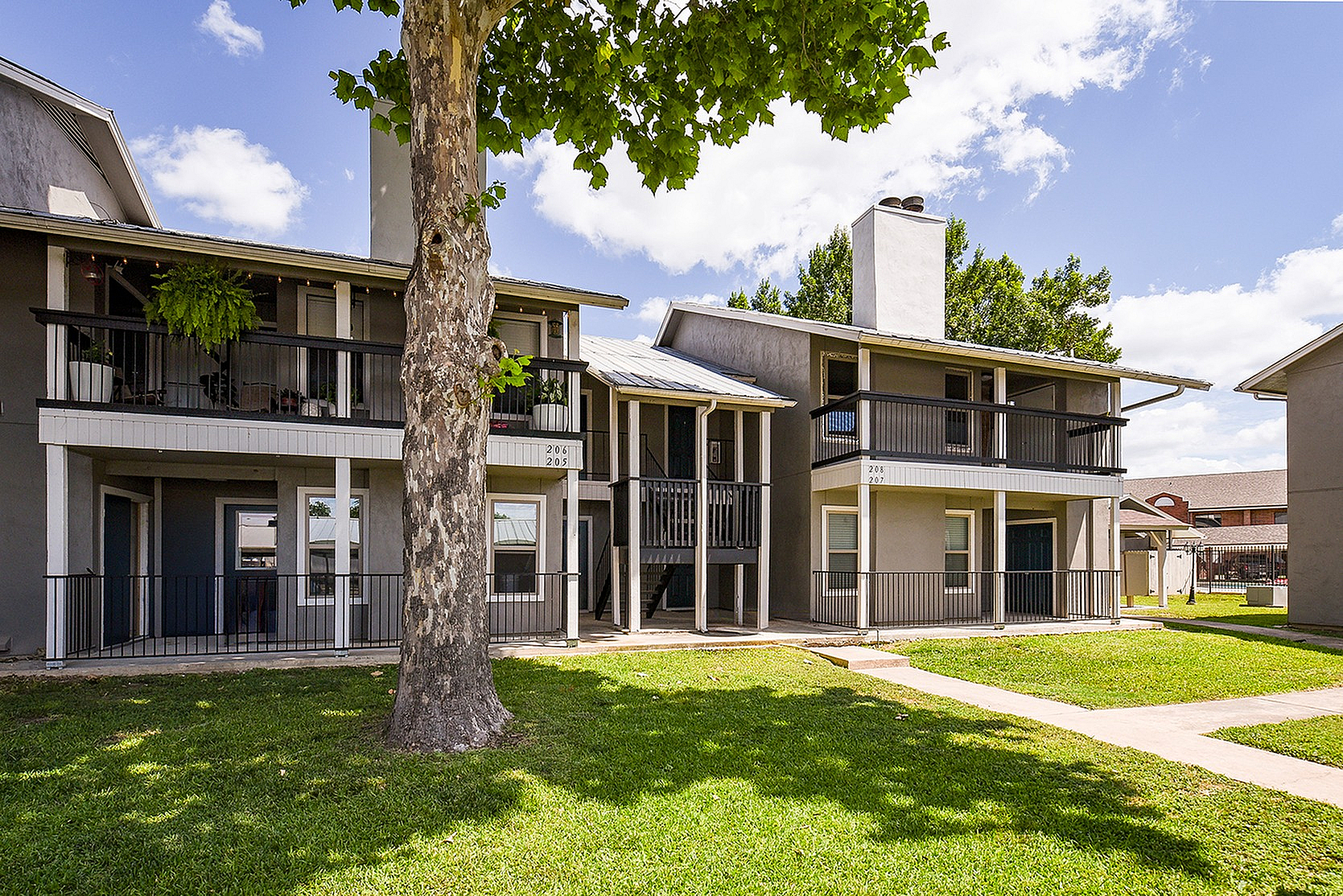 Charming two-story apartment complex with individual balconies, nestled in a serene setting with lush greenery and bright, sunny skies.