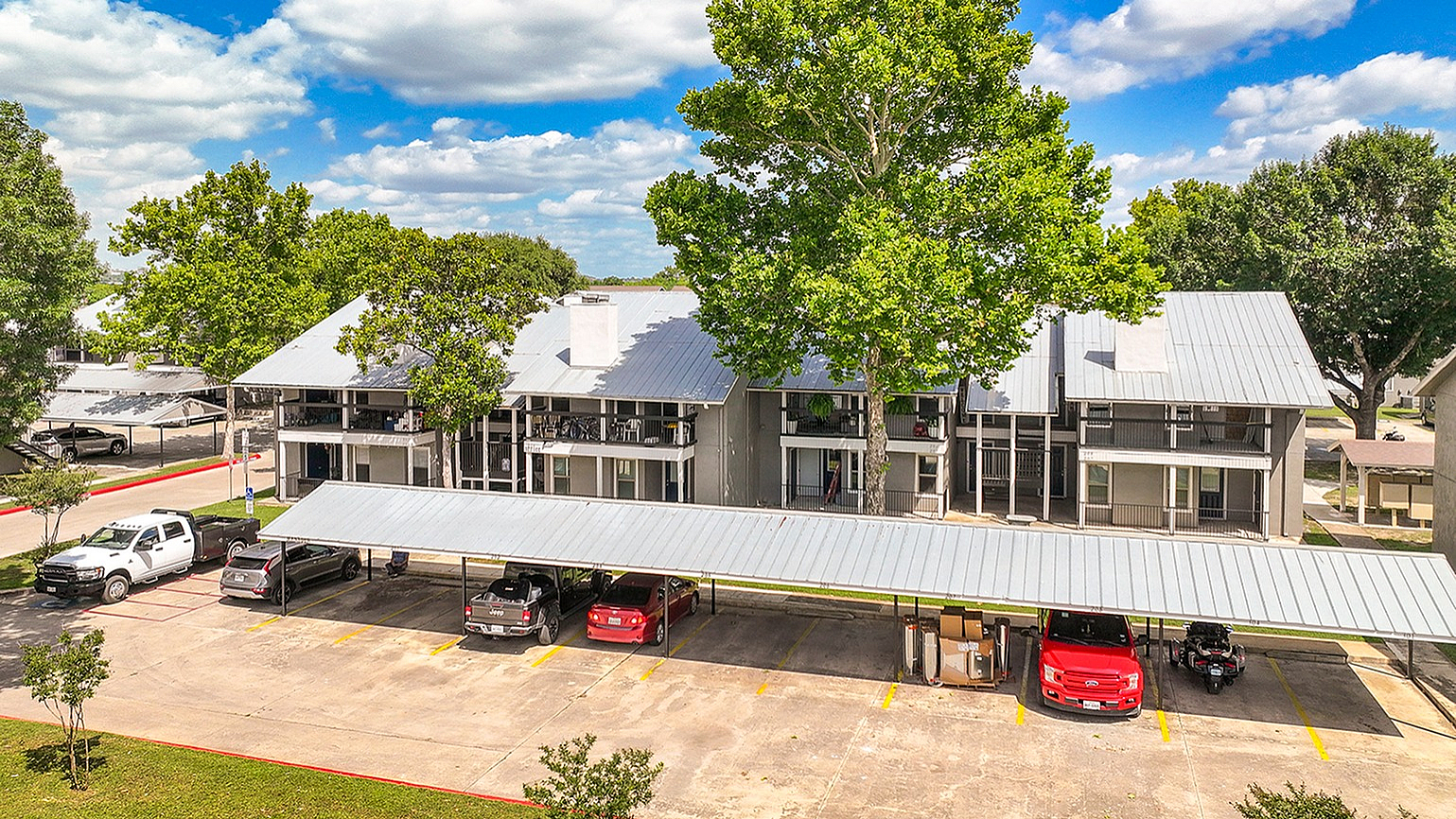 Charming apartment complex with modern architecture, abundant greenery, and covered parking under a bright blue sky.