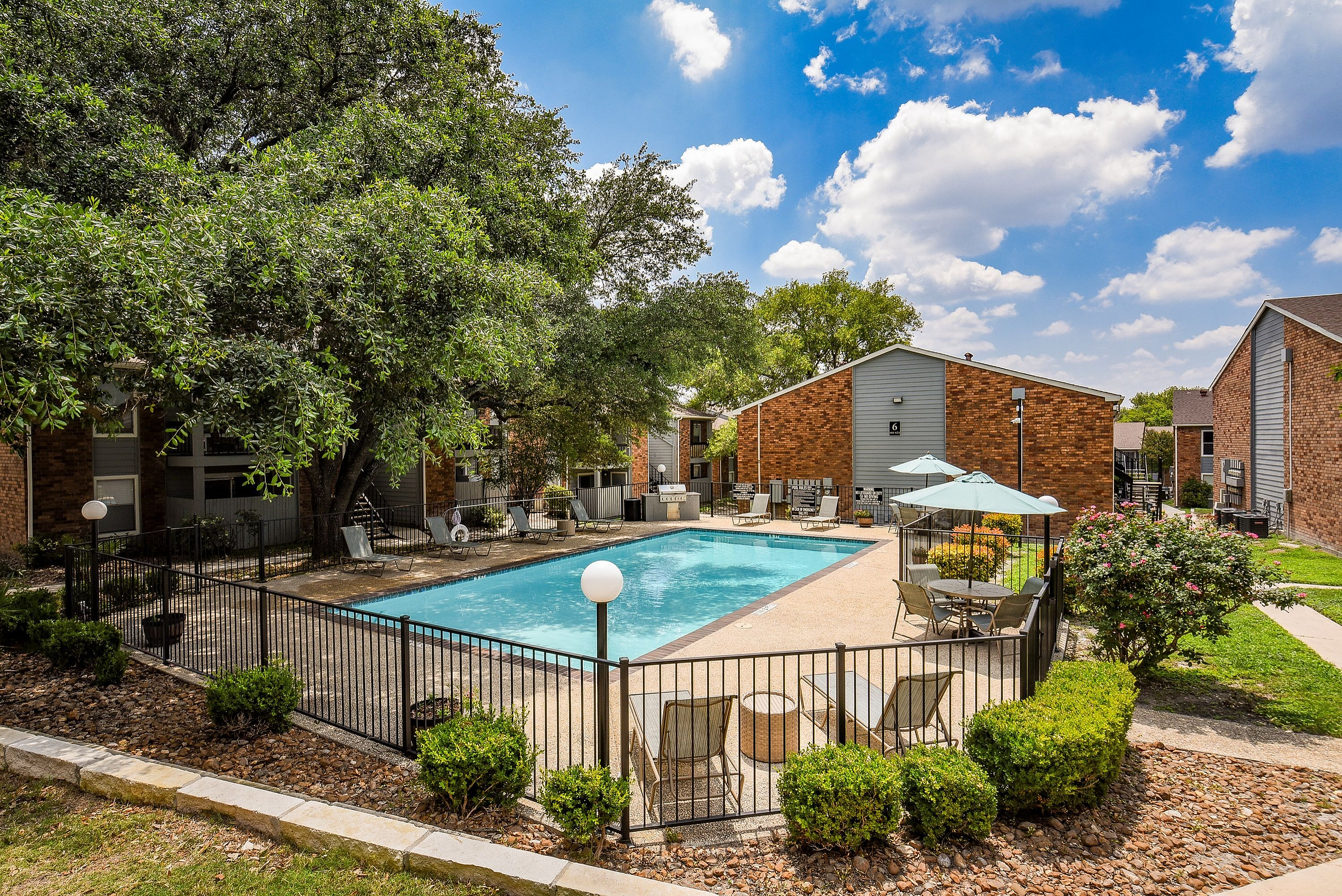 Beautiful apartment complex courtyard featuring a sparkling swimming pool, lush greenery, and stylish lounge chairs under a clear blue sky.