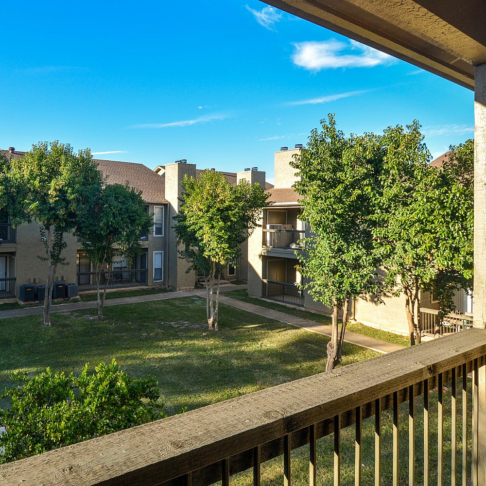 Sunlit apartment verandah overlooking a lush green courtyard with picnic benches and neatly lined trees.