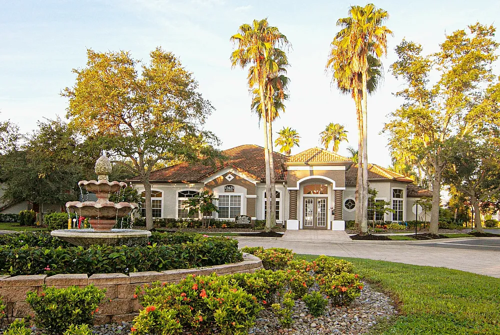 Elegant apartment entrance surrounded by lush greenery, vibrant flowers, and towering palm trees, featuring a charming stone fountain.