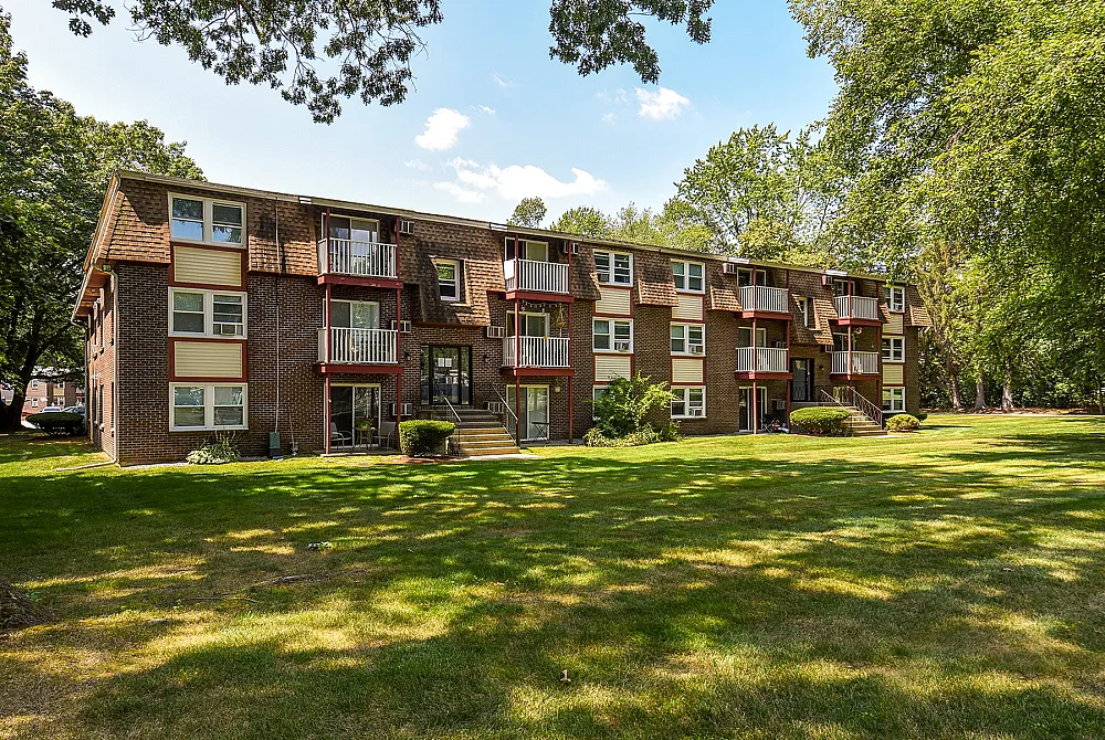 Charming three-story brick apartment building with spacious balconies, surrounded by lush green lawns and tall trees, offering serene living.