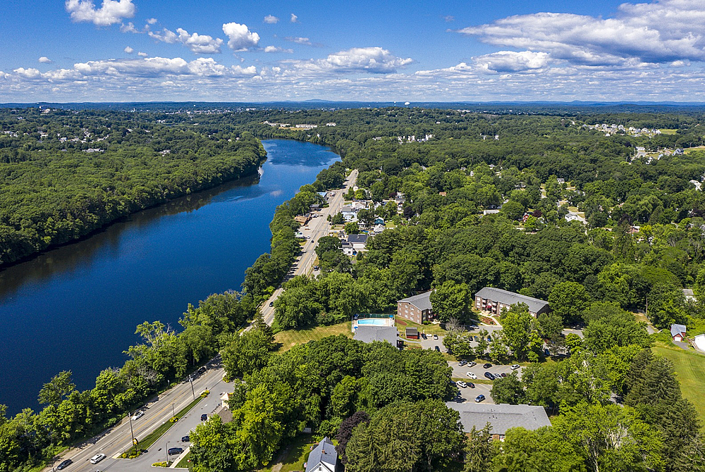 Stunning aerial view showcasing lush greenery along a tranquil riverbank, adjacent to a charming neighborhood with spacious homes and a bright blue swimming pool.