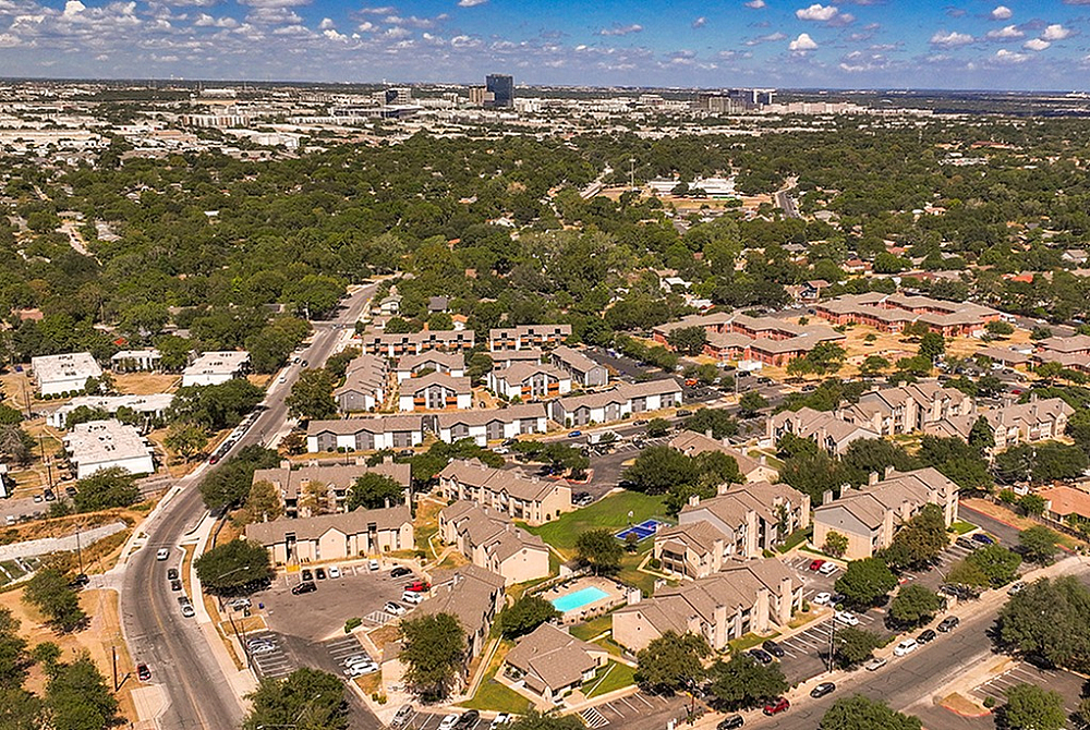 Stunning aerial view of a vibrant residential community surrounded by lush greenery and blue skies, featuring modern townhouses with ample parking and a sparkling swimming pool.