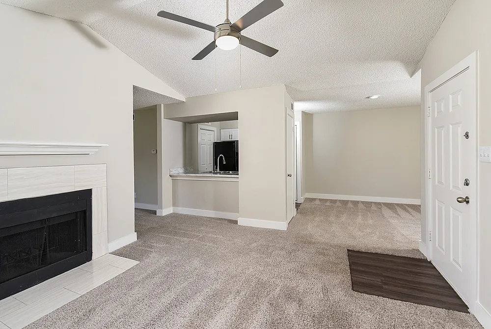 Charming living room with a cozy fireplace, plush carpet, and modern ceiling fan, leading to an open kitchen with sleek appliances.