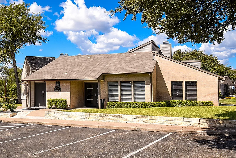 Charming brick apartment building with a quaint entrance, surrounded by lush greenery and a sunny parking area.