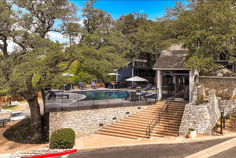 Elegant apartment complex surrounded by lush greenery and featuring a luxurious outdoor pool deck with ample seating and umbrellas, inviting residents to relax and unwind.