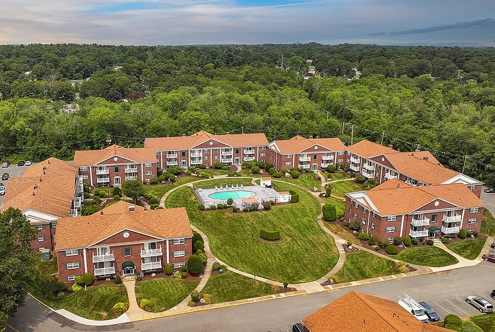 Charming apartment complex with elegant red brick buildings, lush green landscaping, and a central swimming pool, offering a serene residential atmosphere.