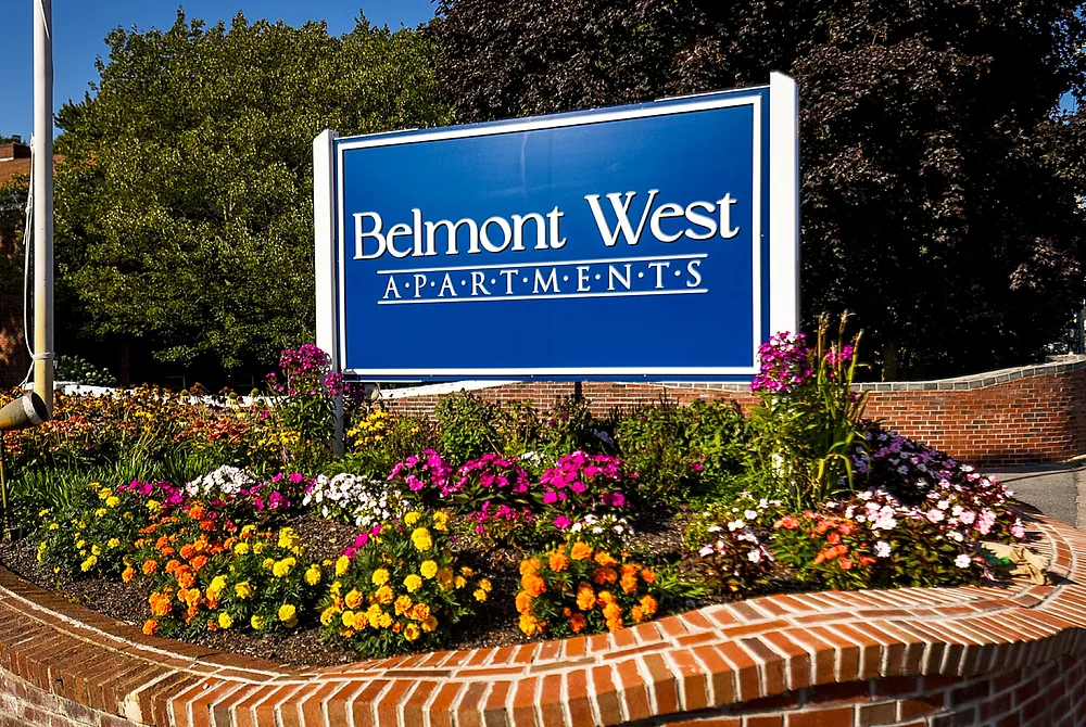 Welcoming entrance sign of Belmont West Apartments surrounded by vibrant flowers and lush greenery.