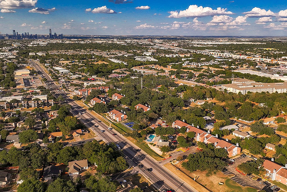 Breathtaking aerial view of a vibrant suburb with lush greenery, modern homes, and distant city skyline under a bright blue sky.