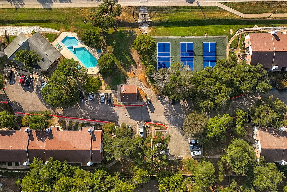 Vibrant aerial view of a luxurious apartment complex with a sparkling swimming pool, multiple tennis courts, and lush green spaces surrounded by mature trees.