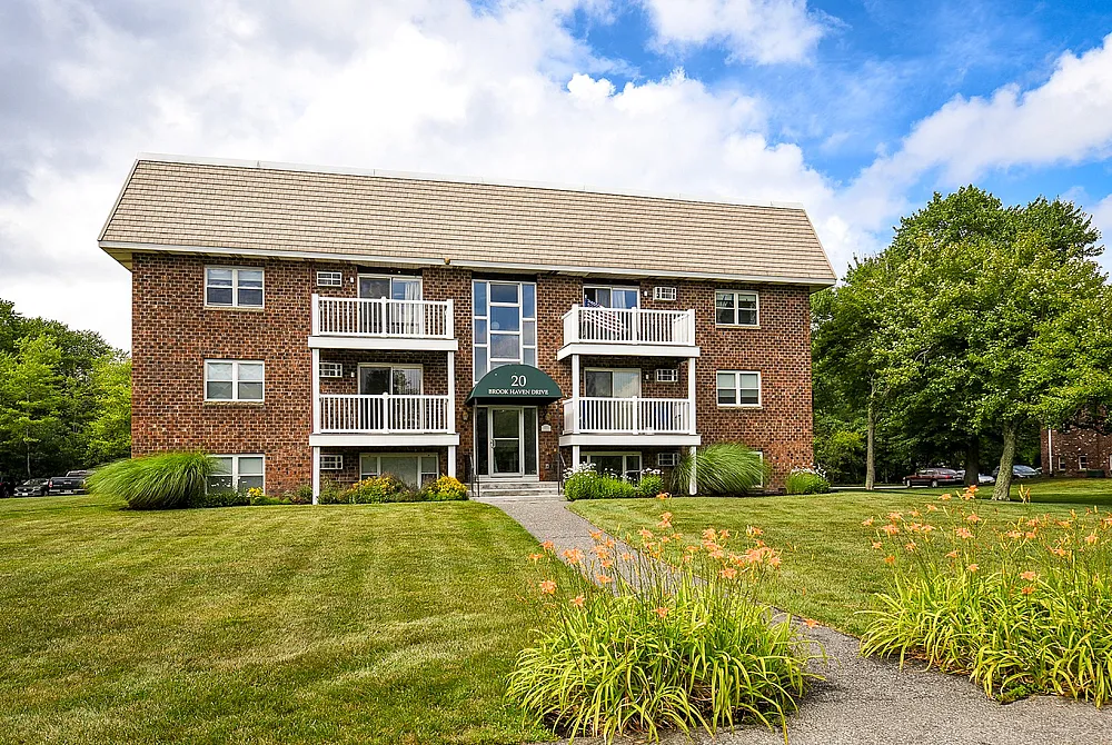 Charming brick apartment building with balconies, surrounded by lush green lawns and vibrant flowers, set against a bright blue sky.