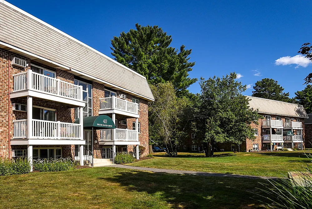Charming brick apartment complex with inviting balconies, nestled in lush green surroundings and under a vibrant blue sky.