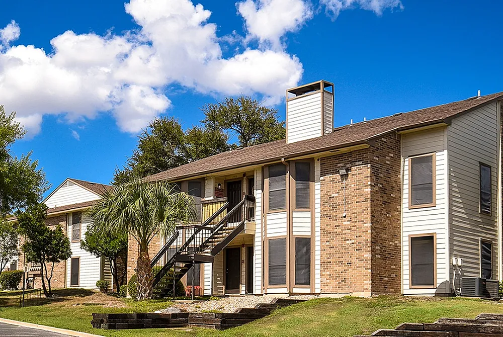 Charming multi-story apartment complex with brick and siding exterior, surrounded by lush greenery and bright blue skies.