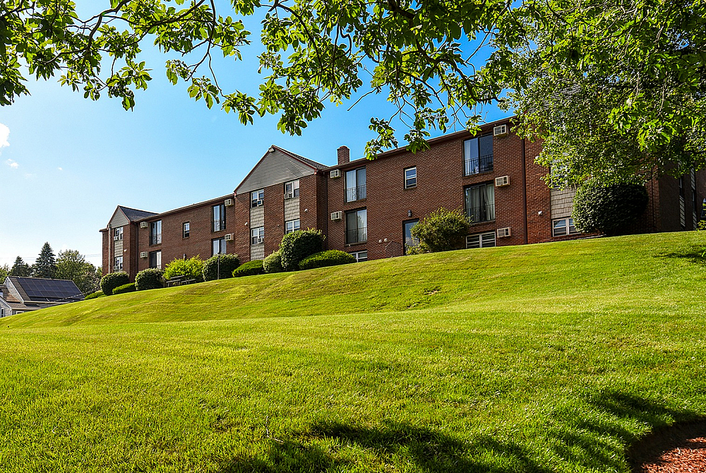 Charming brick apartment complex with lush green lawn and scenic tree-lined surroundings under a clear blue sky.