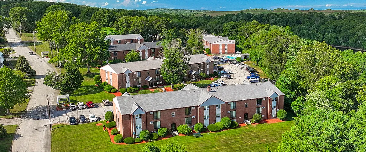 Aerial view of charming brick apartment complex surrounded by lush greenery, featuring well-maintained lawns and ample parking, set against a backdrop of vibrant blue skies.