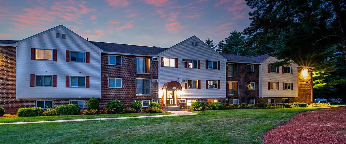Charming apartment building at sunset surrounded by lush greenery and picturesque landscaping. Illuminated entrance exudes warmth and a welcoming atmosphere.