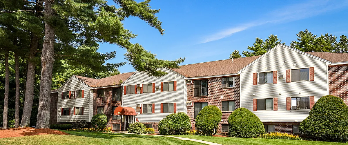 Charming apartment building with brick facade, surrounded by lush greenery and a spacious parking area under a clear blue sky.