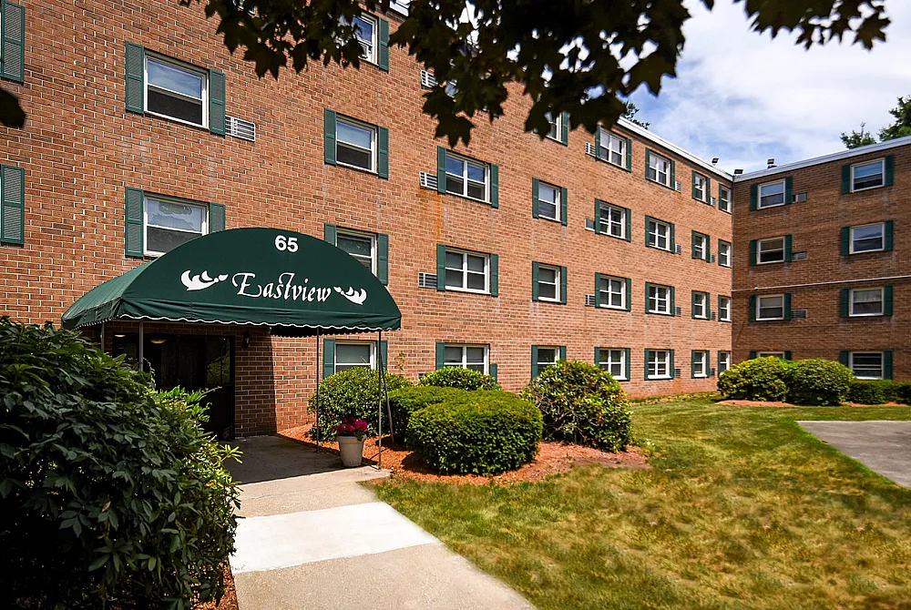 Charming brick apartment building with green accents, lush landscaping, welcoming entrance, and outdoor greenery.