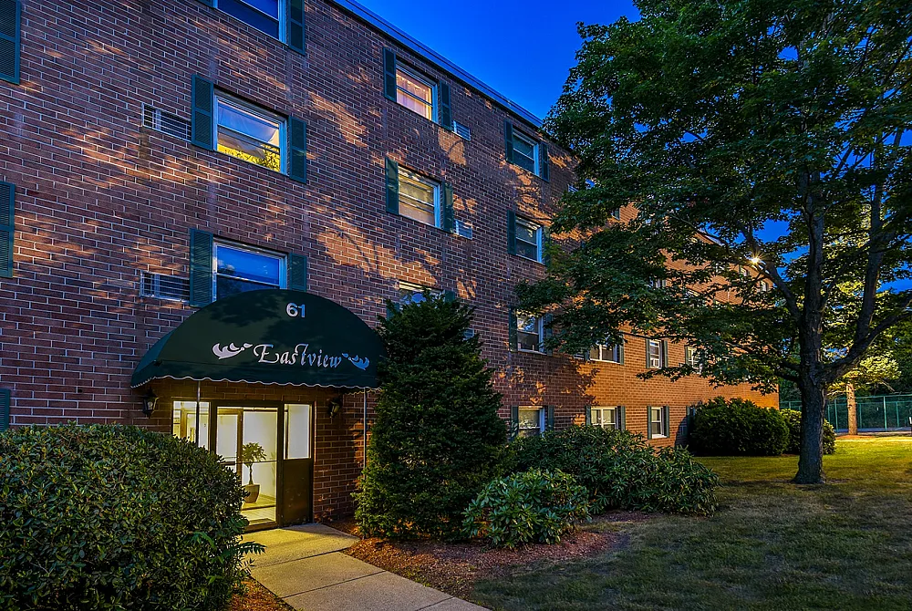 Charming red-brick apartment building with lush greenery, featuring a welcoming entrance canopy under a serene evening sky.